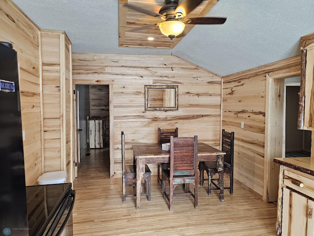 dining space featuring a textured ceiling, vaulted ceiling, light hardwood / wood-style flooring, wooden walls, and ceiling fan