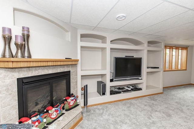 living room featuring built in shelves, carpet flooring, a tiled fireplace, and a drop ceiling