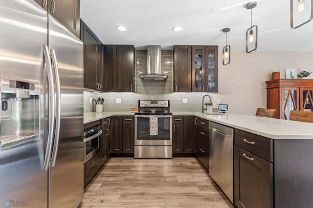 kitchen with light hardwood / wood-style floors, kitchen peninsula, wall chimney range hood, appliances with stainless steel finishes, and decorative light fixtures