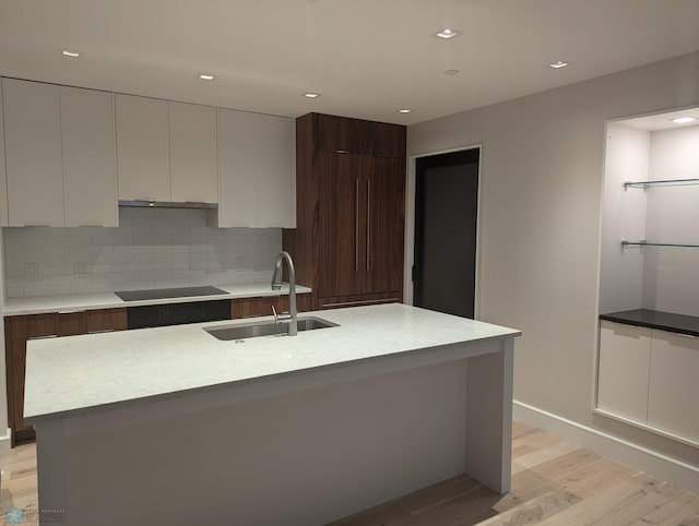 kitchen with sink, a kitchen island with sink, backsplash, black electric cooktop, and light hardwood / wood-style floors