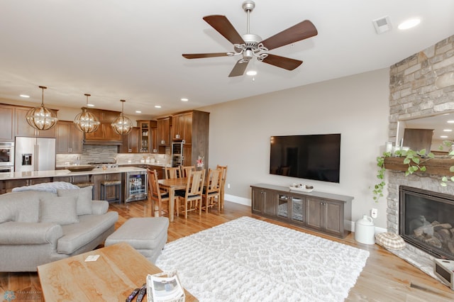 living room featuring light hardwood / wood-style floors, a stone fireplace, ceiling fan with notable chandelier, and beverage cooler