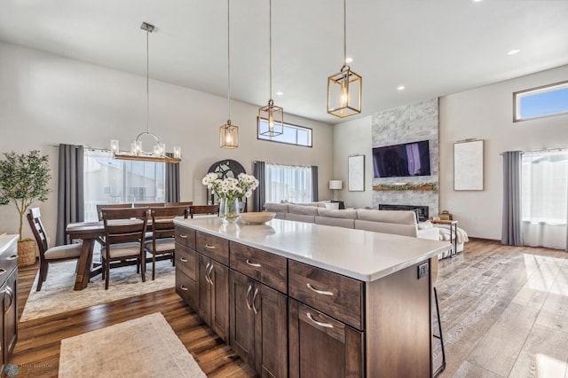 kitchen featuring a towering ceiling, a stone fireplace, dark brown cabinets, dark hardwood / wood-style flooring, and hanging light fixtures