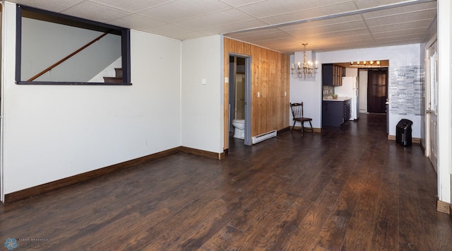 empty room featuring wooden walls, a baseboard heating unit, a drop ceiling, a notable chandelier, and dark hardwood / wood-style flooring