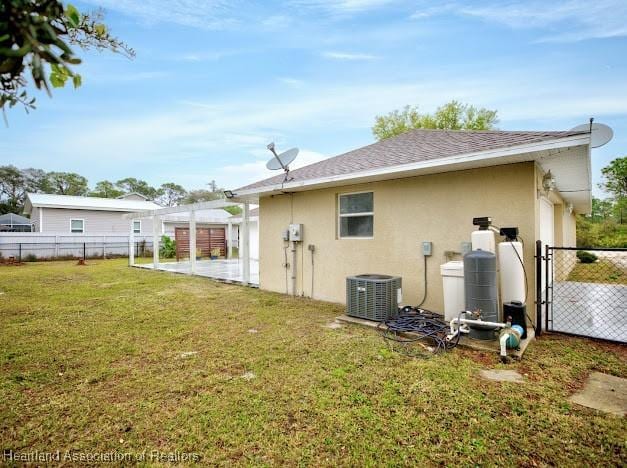 rear view of house with central AC unit, fence, roof with shingles, stucco siding, and a lawn