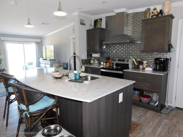 kitchen featuring stainless steel electric stove, a kitchen island with sink, sink, and wall chimney range hood