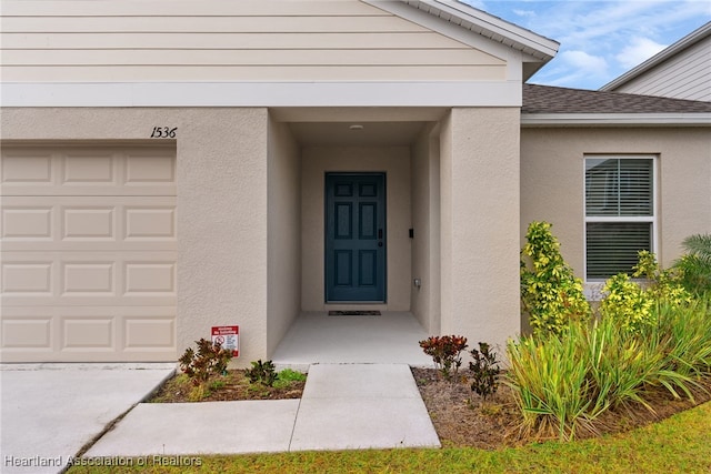 view of exterior entry with a garage, roof with shingles, and stucco siding