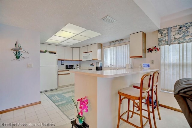 kitchen featuring white appliances, a textured ceiling, cream cabinetry, a kitchen bar, and kitchen peninsula