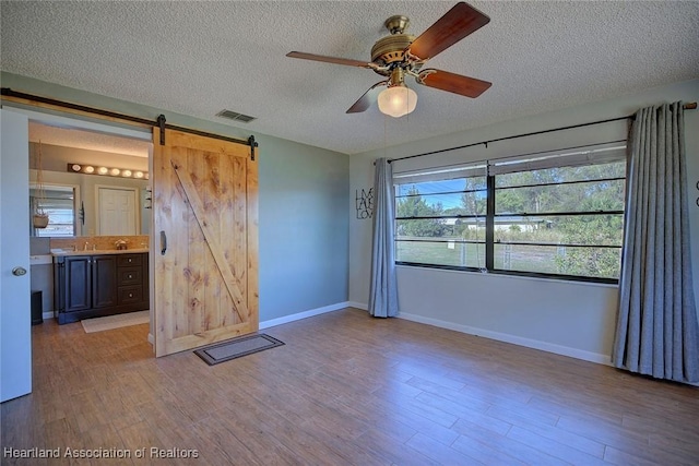 unfurnished bedroom featuring ensuite bath, ceiling fan, a barn door, light hardwood / wood-style flooring, and a textured ceiling