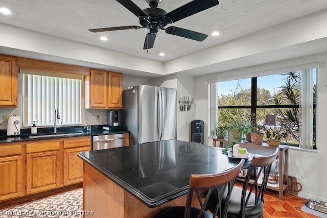 kitchen featuring stainless steel appliances, dark countertops, brown cabinetry, a sink, and a kitchen island