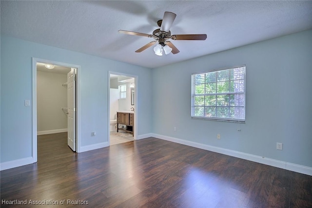 unfurnished bedroom featuring a ceiling fan, baseboards, a spacious closet, ensuite bath, and dark wood-type flooring