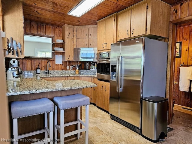 kitchen with kitchen peninsula, light stone countertops, stainless steel appliances, wooden ceiling, and wood walls