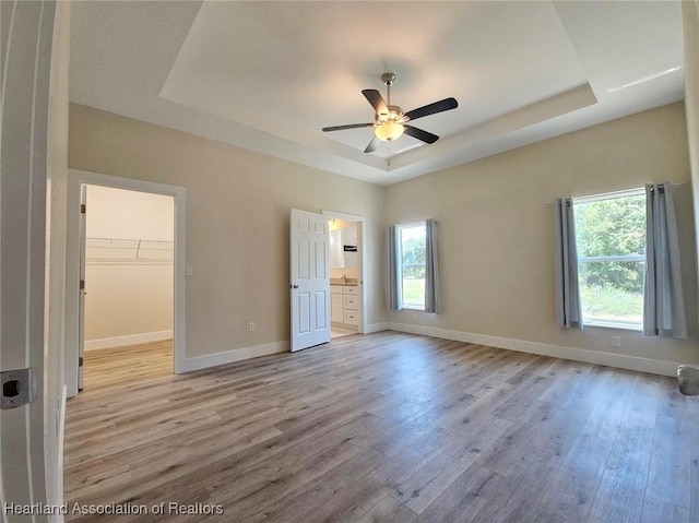 unfurnished bedroom featuring ensuite bathroom, light wood-type flooring, a spacious closet, a raised ceiling, and baseboards