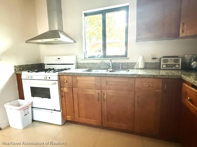 kitchen with sink, dark stone counters, light tile patterned floors, white gas range oven, and wall chimney range hood