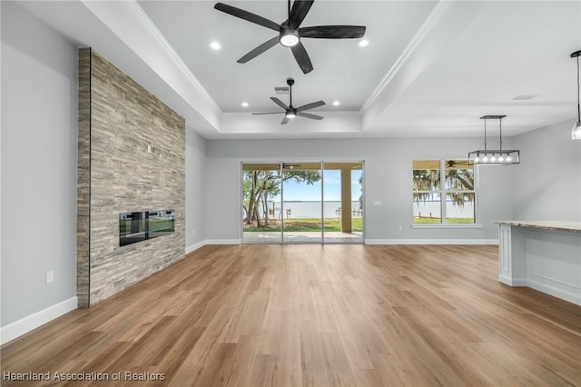 unfurnished living room featuring ceiling fan, a raised ceiling, a stone fireplace, crown molding, and light wood-type flooring