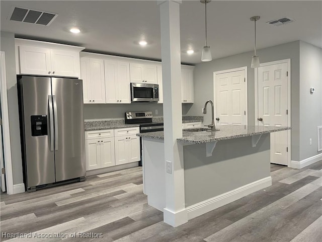 kitchen with light stone countertops, appliances with stainless steel finishes, a center island with sink, decorative light fixtures, and white cabinetry