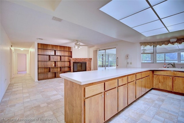 kitchen with a sink, a peninsula, light countertops, a brick fireplace, and ceiling fan