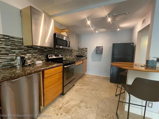 kitchen with sink, rail lighting, stainless steel appliances, tasteful backsplash, and dark stone counters