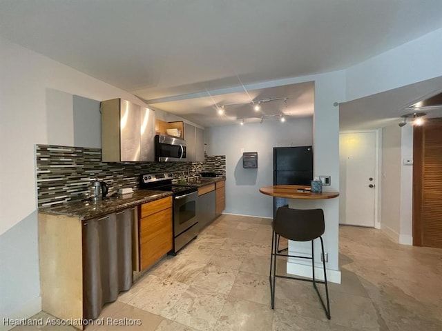 kitchen featuring rail lighting, stainless steel appliances, tasteful backsplash, dark stone counters, and a kitchen bar