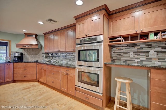 kitchen featuring stainless steel double oven, light wood-style flooring, visible vents, dark stone counters, and custom range hood