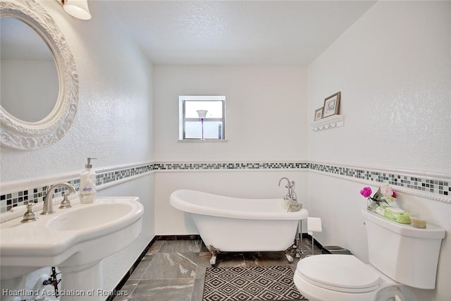 bathroom with a textured wall, toilet, marble finish floor, a freestanding tub, and a sink