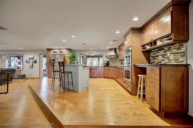 kitchen featuring custom range hood, a kitchen breakfast bar, stainless steel appliances, light wood-type flooring, and backsplash