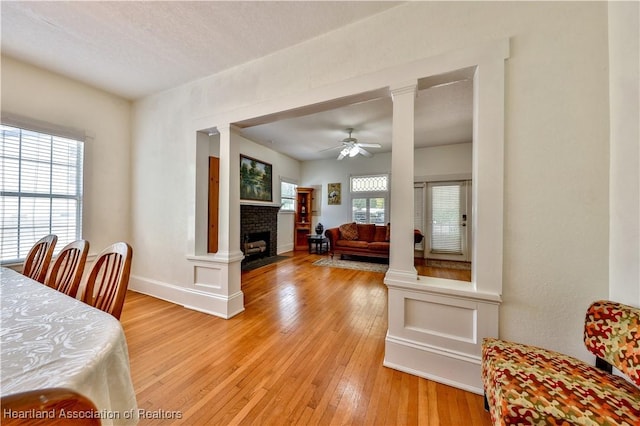 dining area featuring ceiling fan, a brick fireplace, decorative columns, light hardwood / wood-style floors, and a textured ceiling