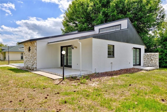 rear view of house featuring a patio, stone siding, stucco siding, a lawn, and french doors