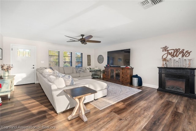 living room featuring dark wood finished floors, a glass covered fireplace, visible vents, and a ceiling fan