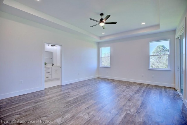 unfurnished bedroom featuring a tray ceiling, ensuite bath, wood finished floors, and baseboards