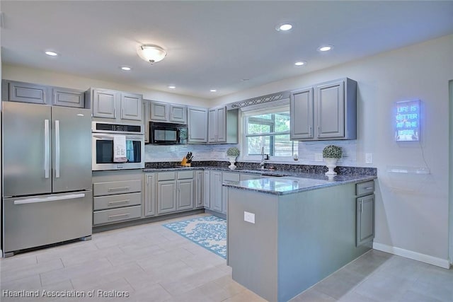 kitchen featuring gray cabinets, backsplash, a peninsula, stainless steel appliances, and dark stone counters