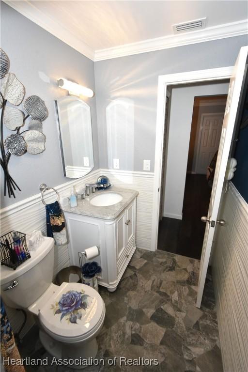 half bathroom featuring visible vents, toilet, ornamental molding, wainscoting, and vanity