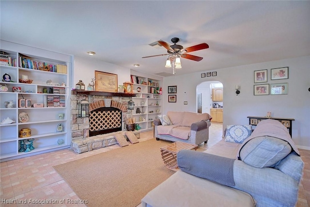 living area with arched walkways, built in shelves, a stone fireplace, and a ceiling fan