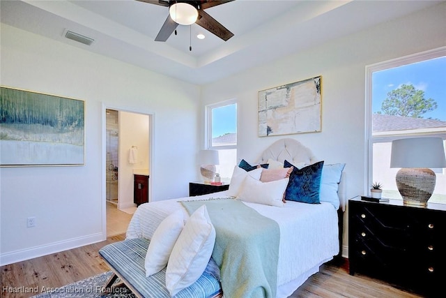 bedroom featuring ceiling fan, ensuite bathroom, a raised ceiling, and light hardwood / wood-style flooring