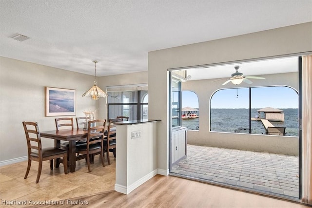 dining space featuring ceiling fan with notable chandelier, a textured ceiling, light hardwood / wood-style flooring, and a water view