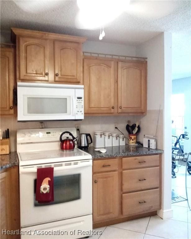 kitchen with light brown cabinetry, dark stone counters, light tile patterned floors, and white appliances