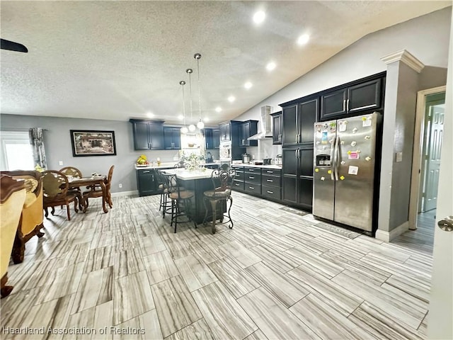 kitchen with stainless steel refrigerator with ice dispenser, a kitchen breakfast bar, wall chimney exhaust hood, pendant lighting, and a kitchen island