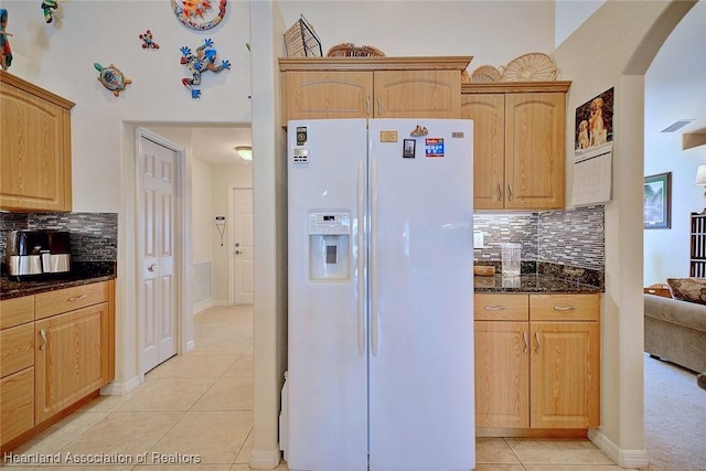 kitchen with decorative backsplash, light brown cabinetry, light tile patterned floors, dark stone countertops, and white fridge with ice dispenser