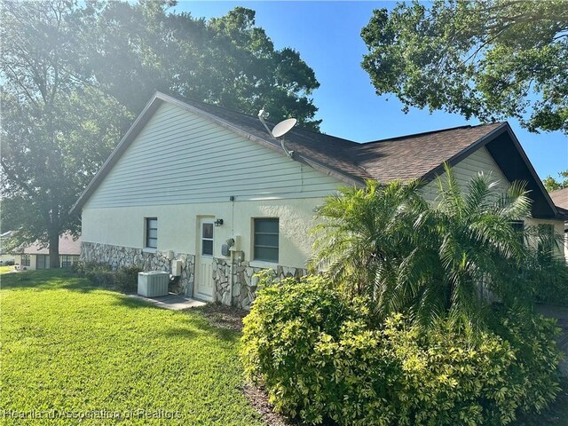 back of property with roof with shingles, a yard, central AC, stucco siding, and stone siding