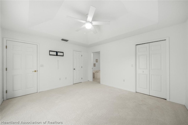 unfurnished bedroom with ceiling fan, a raised ceiling, light carpet, and multiple closets