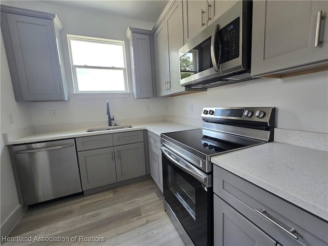 kitchen with appliances with stainless steel finishes, light stone counters, gray cabinetry, sink, and light hardwood / wood-style floors