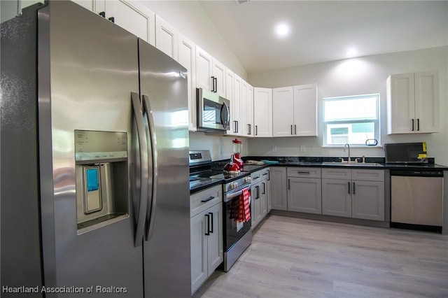 kitchen with light wood finished floors, lofted ceiling, a sink, appliances with stainless steel finishes, and dark countertops