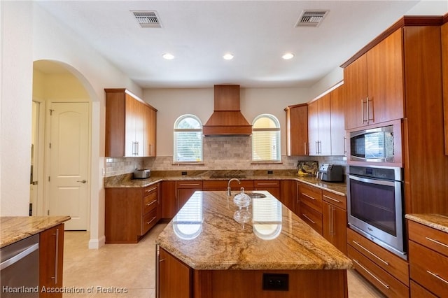 kitchen with decorative backsplash, light stone counters, custom exhaust hood, stainless steel appliances, and a center island with sink