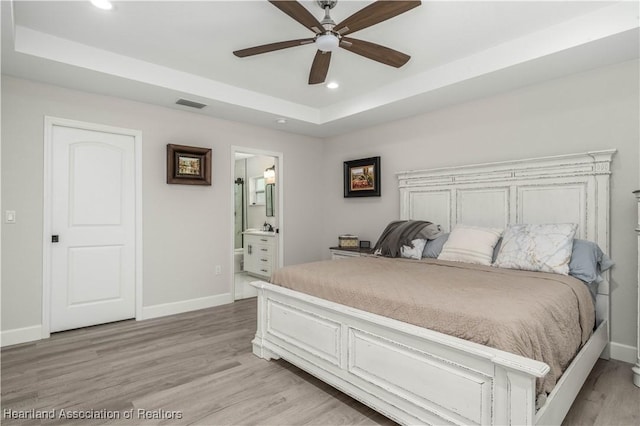 bedroom with a tray ceiling, ensuite bath, ceiling fan, and light hardwood / wood-style flooring