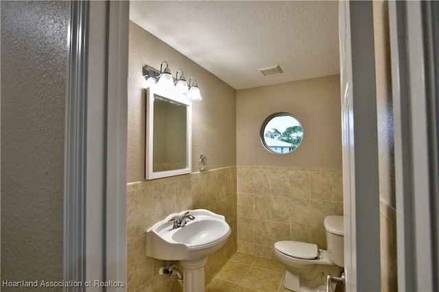 half bathroom with tile patterned flooring, visible vents, tile walls, toilet, and a textured ceiling