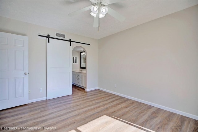 unfurnished bedroom with ensuite bathroom, sink, light hardwood / wood-style flooring, ceiling fan, and a barn door