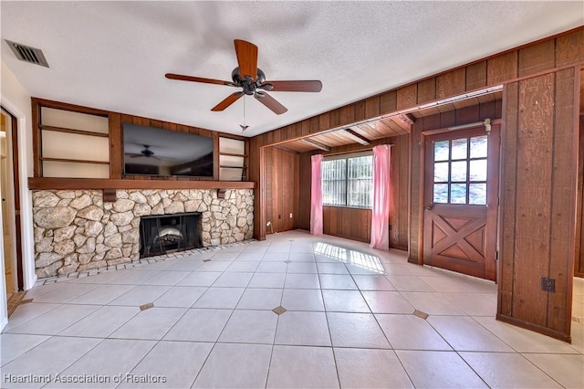 unfurnished living room featuring a textured ceiling, ceiling fan, wooden walls, light tile patterned floors, and a stone fireplace