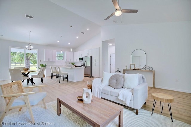 living area featuring high vaulted ceiling, light wood-style flooring, baseboards, ceiling fan with notable chandelier, and visible vents