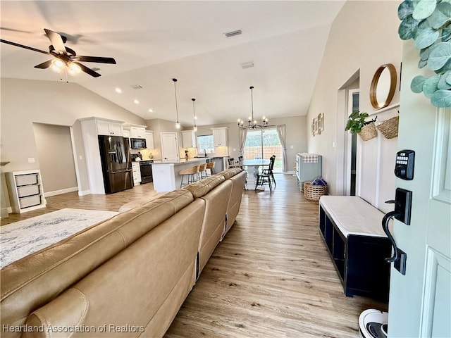 living area featuring ceiling fan with notable chandelier, wood finished floors, visible vents, baseboards, and vaulted ceiling