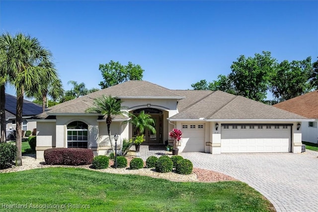 ranch-style house featuring a front yard, an attached garage, a shingled roof, stucco siding, and decorative driveway