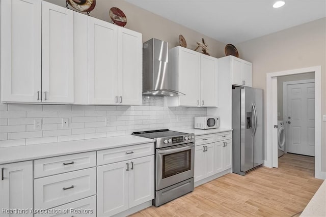 kitchen with washer / dryer, white cabinetry, light wood-type flooring, appliances with stainless steel finishes, and wall chimney range hood